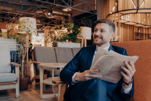 Jovem empresário confiante lendo jornal e as últimas notícias financeiras enquanto está sentado na poltrona no interior de uma cafeteria moderna, enquanto espera que um colega de trabalho tome o café da manhã juntos