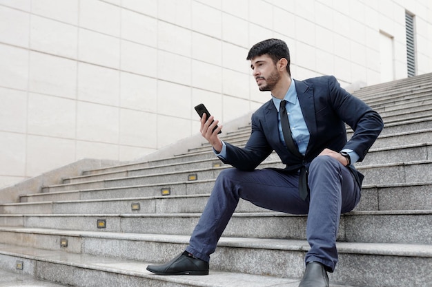 Jovem empresário confiante sentado em degraus ao ar livre e lendo artigo on-line no smartphone