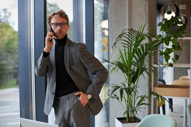 Jovem empresário confiante usando telefone negociando com investidores aguardando janela no escritório