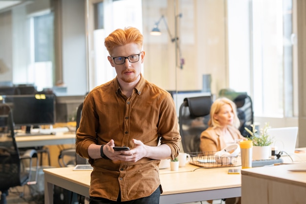 Jovem empresário contemporâneo de óculos e roupa casual inteligente, usando o telefone celular e olhando para você enquanto está de pé na mesa no escritório