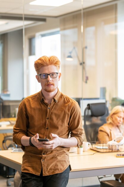 Jovem empresário contemporâneo e confiante usando óculos, usando o telefone celular e olhando para você enquanto está perto da mesa