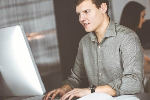 Jovem empresário de cabelos escuros e programador de camisa verde está trabalhando duro em seu computador, enquanto está sentado à mesa em um gabinete moderno com uma colega no fundo. Conceito de sucesso