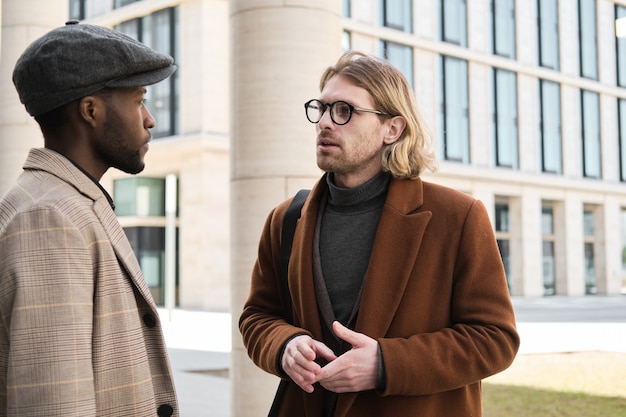 Jovem empresário de casaco e óculos conversando com seu colega na rua