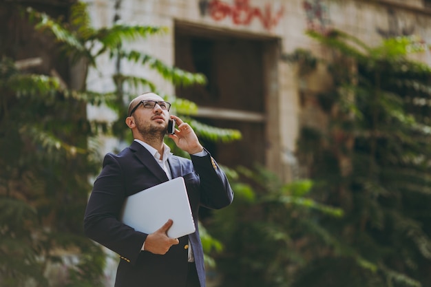 Foto jovem empresário de sucesso em camisa branca, terno clássico, óculos. homem de pé com o computador laptop pc, falando no telefone perto de ruínas, destroços, edifício de pedra ao ar livre. escritório móvel, conceito de negócio.