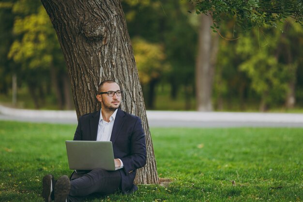 Jovem empresário de sucesso em camisa branca, terno clássico, óculos. Homem sentar na grama, trabalhar no computador laptop pc no parque da cidade, no gramado verde ao ar livre na natureza. Escritório móvel, conceito de negócio.