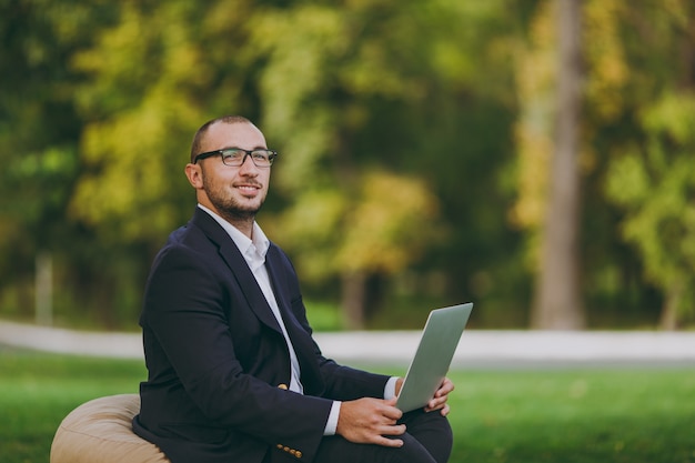 Jovem empresário de sucesso em camisa branca, terno clássico, óculos. Homem sente-se no pufe macio, trabalhando no computador laptop pc no parque da cidade, no gramado verde ao ar livre na natureza. Escritório móvel, conceito de negócio.