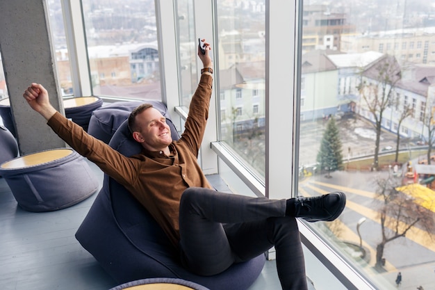Jovem empresário de sucesso está sentado perto de uma grande janela no escritório e relaxando. Descanse depois de um dia de trabalho