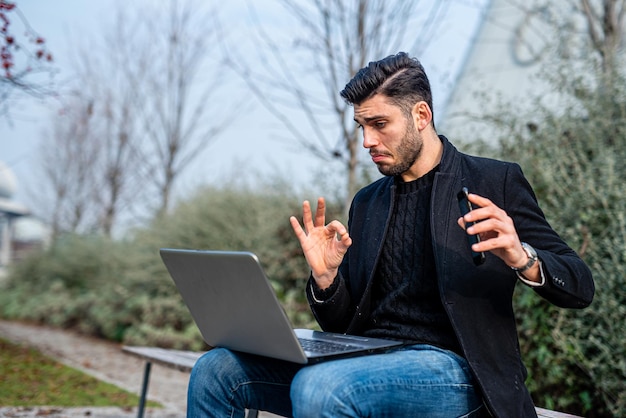 Jovem empresário de terno casual trabalhando em teletrabalho empresário masculino sentado em um banco de parque público falando com colegas em videochamada homem positivo e bem sucedido