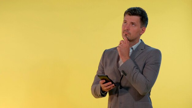 Foto jovem empresário de terno olhando no smartphone e pensando em algo sobre fundo amarelo ele ...