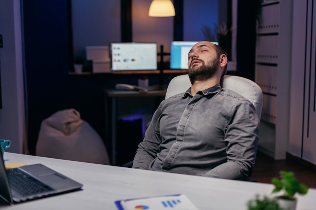 Jovem empresário, descansando na cadeira enquanto trabalhava no prazo. Funcionário workaholic adormecendo por causa de enquanto trabalhava sozinho tarde da noite no escritório para um projeto importante da empresa.
