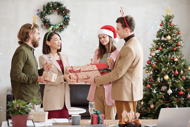 Jovem empresário elegante com bebida e presente de Natal embalado para uma das colegas enquanto a parabeniza na festa no escritório