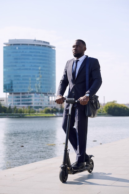 Jovem empresário elegante em pé na scooter elétrica à beira do rio contra um edifício moderno e alto, indo para o trabalho pela manhã