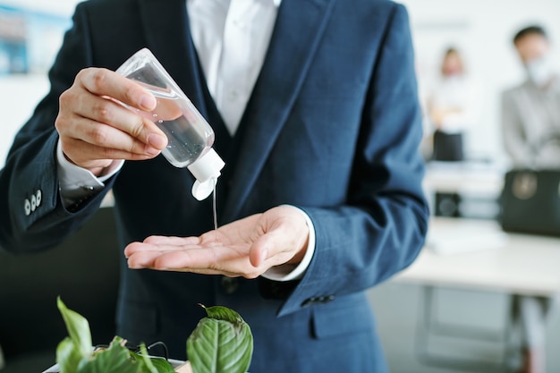 Jovem empresário elegante em terno pingando desinfetante de uma garrafa de plástico nas mãos no início do dia de trabalho no escritório