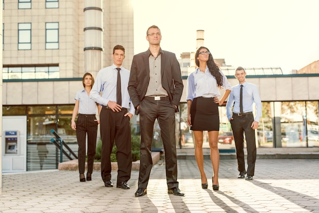 Jovem empresário, elegantemente vestido com as mãos nos bolsos, de pé orgulhosamente com sua equipe de jovens empresárias e empresários na frente do prédio iluminado sol iluminado.