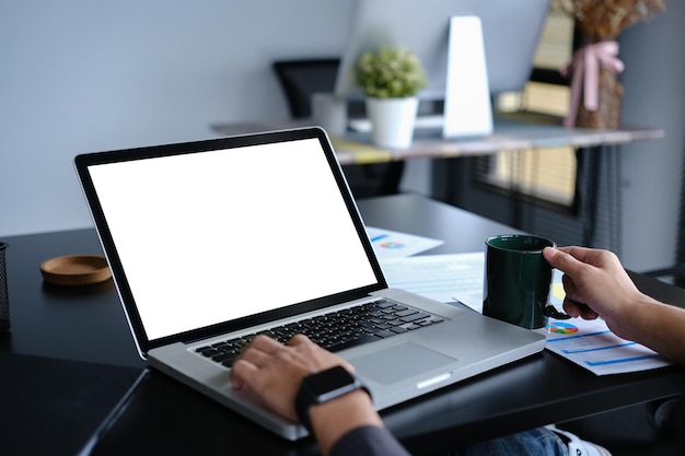 Jovem empresário em close-up segurando uma xícara de café e usando um computador portátil em uma mesa de escritório preta