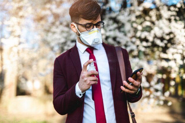 Jovem empresário em pé no parque, usando máscara protetora e desinfetando o telefone inteligente com álcool.