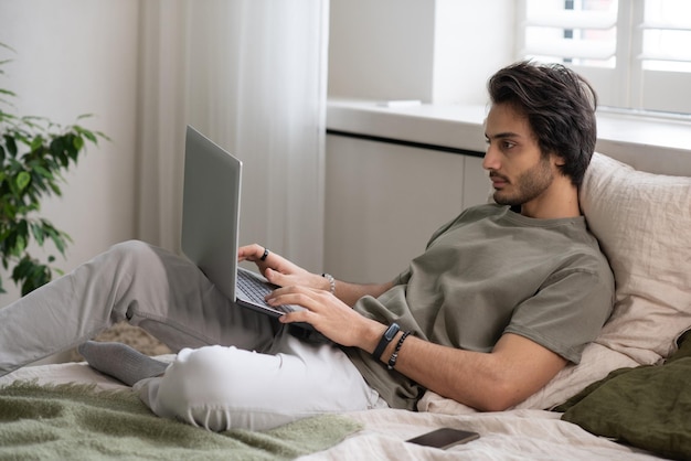 Jovem empresário em roupas casuais, deitado na cama e digitando no teclado do laptop