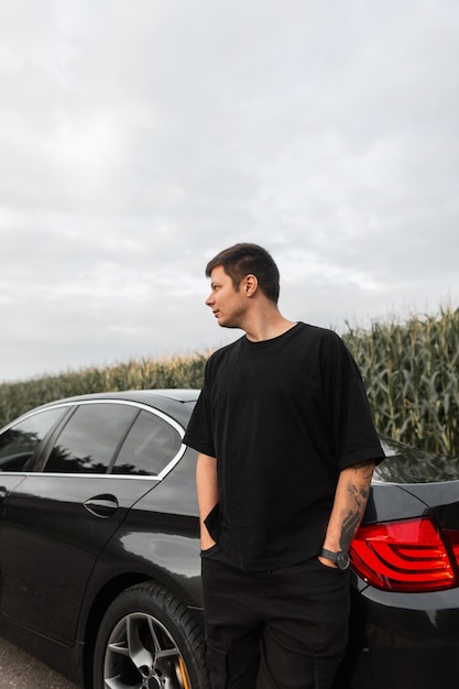 Jovem empresário em uma camiseta preta com uma tatuagem no braço ao lado de um carro preto na estrada fora da cidade Viajar de carro