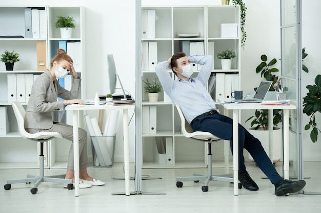 Jovem empresário em uma máscara protetora, fazendo um brainstorming na frente do laptop enquanto sua colega pensa durante o trabalho em um novo projeto