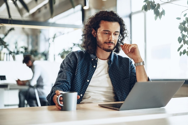 Jovem empresário está usando laptop e segurando uma xícara de café no escritório