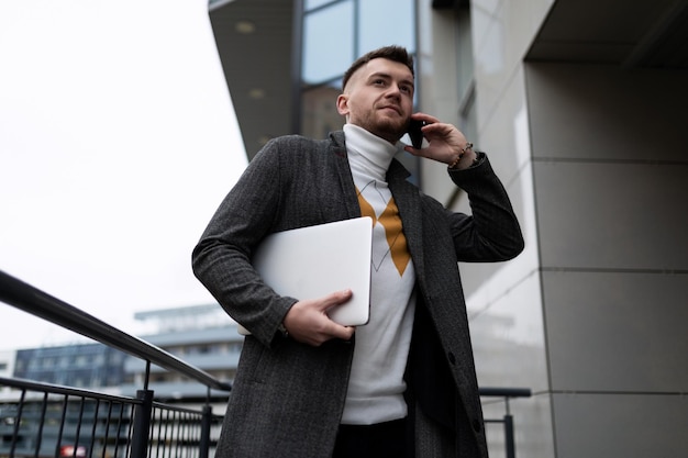 Jovem empresário falando em um telefone celular com um laptop nas mãos ao lado da entrada do