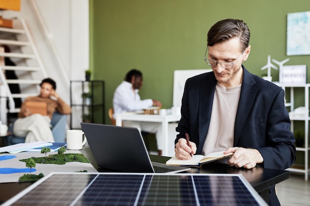 Jovem empresário fazendo anotações no caderno enquanto trabalhava na mesa com laptop e painel solar