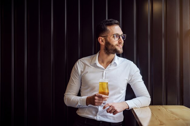 Jovem empresário geeky sorridente, encostado na mesa ao lado da janela e olhando através dela, segurando um copo de cerveja e relaxando depois do trabalho.