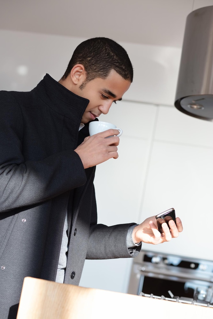 Foto jovem empresário gosta de café e verifica seu telefone em uma cozinha moderna equilibrando trabalho e lazer
