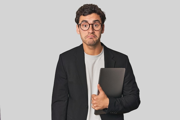 Jovem empresário hispânico com laptop encolhe de ombros e olhos abertos confuso