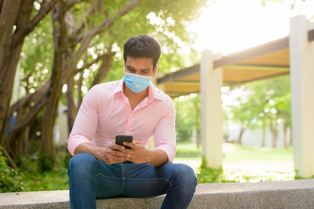 Jovem empresário indiano com máscara usando telefone e sentado no parque