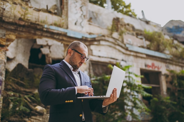 Jovem empresário inteligente de sucesso em camisa branca, terno clássico, óculos. Homem de pé e trabalhando no computador laptop pc perto de ruínas, detritos, construção de pedra ao ar livre. Escritório móvel, conceito de negócio.