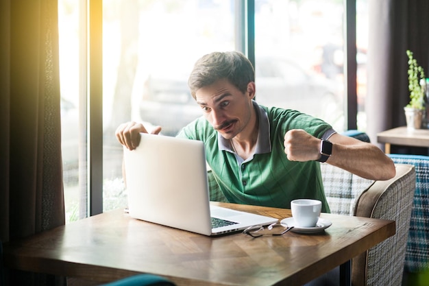 Jovem empresário irritado em camiseta verde sentado com cara agressiva e punhos de boxe e tentar socar seu laptop. problema no conceito de negócios e freelancer. interior, perto de uma grande janela durante o dia.