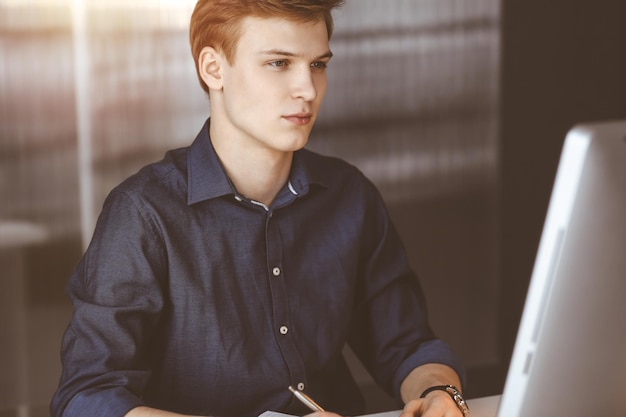 Jovem empresário loiro pensando em estratégia em seu local de trabalho com computador em um escritório escuro, brilho de luz no fundo