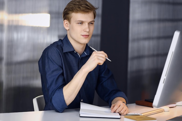 Jovem empresário loiro pensando em estratégia em seu local de trabalho com computador. Startup business significa trabalhar duro e sem tempo para alcançar o sucesso.