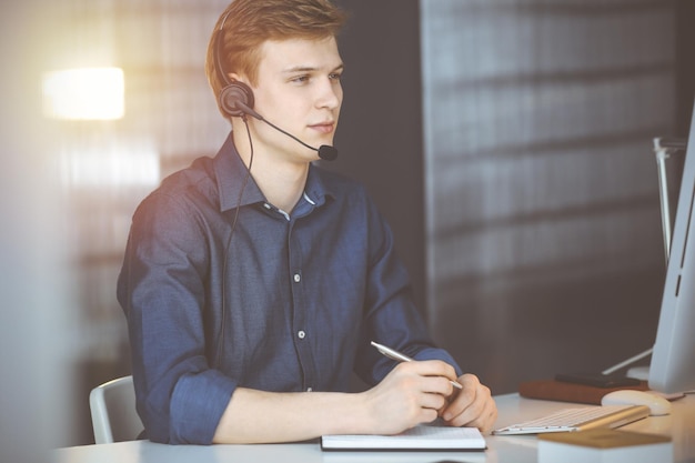 Jovem empresário loiro usando fone de ouvido e computador em um escritório escuro, brilho de luz no fundo. Startup de negócios significa trabalhar duro.