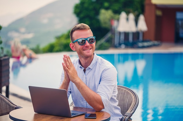 Jovem empresário masculino em óculos de sol trabalha em um laptop sentado em uma mesa perto da piscina Trabalho remoto Freelancer