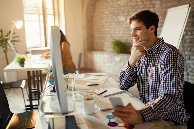 Jovem empresário navegando na net no PC desktop enquanto usa o celular no escritório