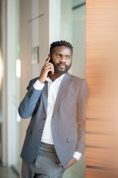 Jovem empresário negro confiante e satisfeito com uma jaqueta em pé na parede e falando ao celular