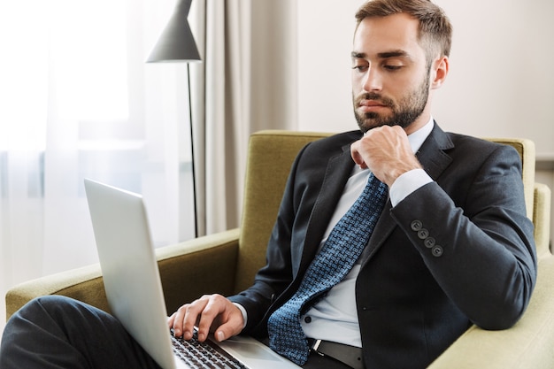 Jovem empresário pensativo atraente vestindo terno, sentado em uma cadeira no quarto do hotel, trabalhando em um laptop