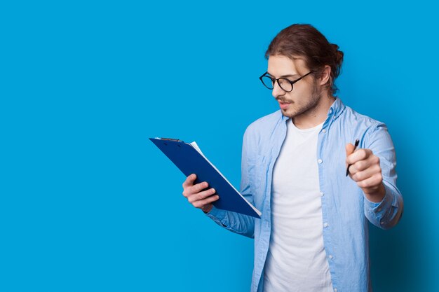 Jovem empresário posando em uma parede azul com espaço livre, olhando em uma pasta e segurando uma caneta