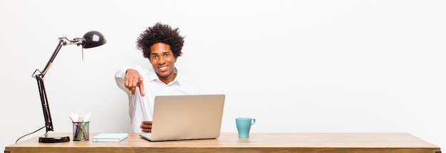 Jovem empresário preto apontando a câmera com um sorriso satisfeito, confiante e amigável, escolhendo você em uma mesa