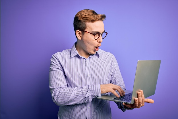 Jovem empresário ruivo bonito trabalhando usando laptop sobre fundo roxo assustado em estado de choque com uma cara de surpresa com medo e animado com expressão de medo