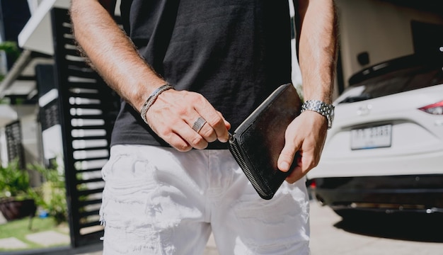 Jovem empresário segurando a carteira nas mãos contra sua casa moderna com garagem e carro