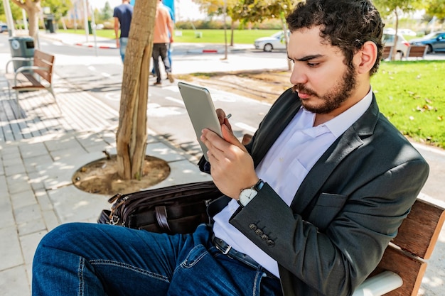 Jovem empresário sentado em um banco no parque segurando seu tablet