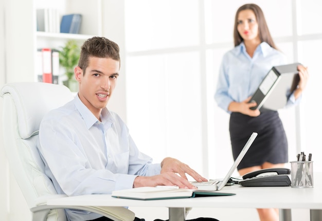 Jovem empresário sentado na cadeira do escritório na frente de um laptop, com um sorriso olhando para a câmera no fundo é uma jovem segurando um fichário.