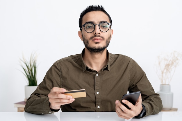 Jovem empresário sério com cartão de crédito e smartphone vai fazer o pagamento das mercadorias que encomendou pela internet