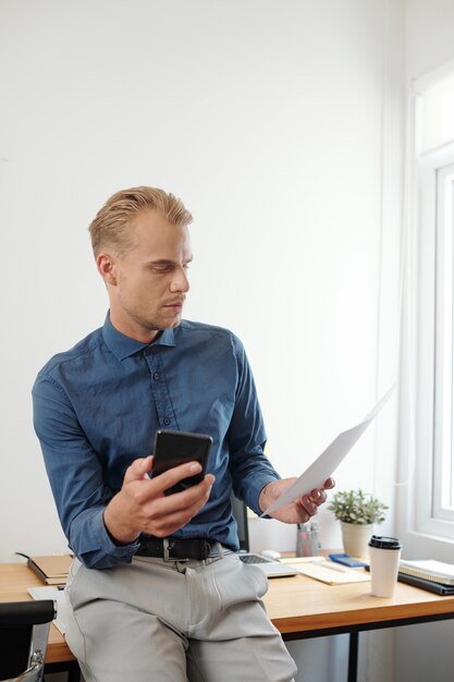 Jovem empresário sério e pensativo com o smartphone na mão, sentado em sua mesa e lendo relatório ou e-mail impresso