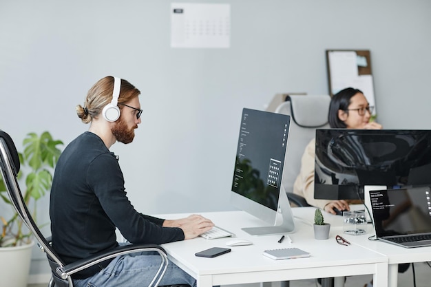 Jovem empresário sério em roupas casuais e fones de ouvido trabalhando na mesa em frente ao monitor do computador no escritório de espaço aberto