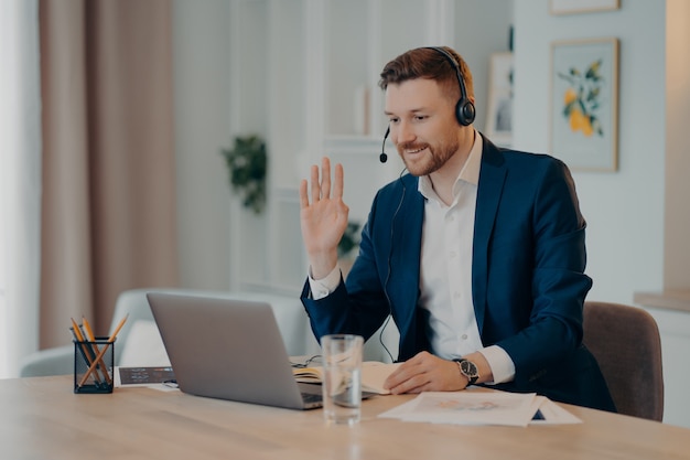 Foto jovem empresário sorridente com fone de ouvido e terno dizendo olá, acenando para alguém e sorrindo enquanto trabalhava remotamente em casa, tendo uma videochamada com colegas ou participando de uma conferência na web