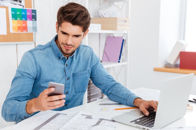 Jovem empresário sorridente de óculos usando laptop e smartphone no local de trabalho